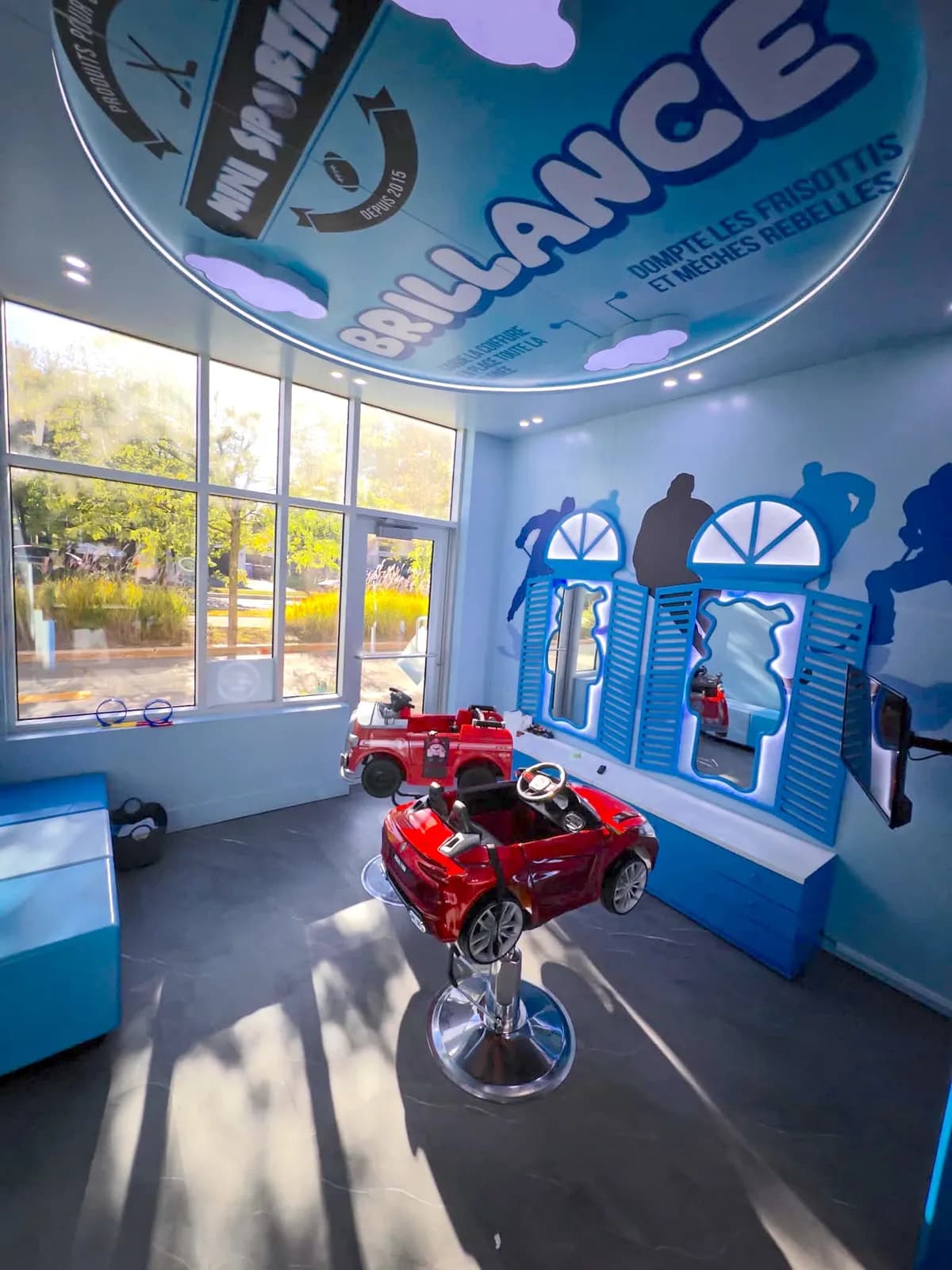 La section enfants d'une succursale Le Barbu Sportif : fauteuils-voitures rouges, miroirs à volets bleus et plafond « Brillance »